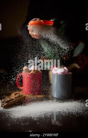 Una persona cosparge zucchero sulle bevande calde di cioccolata in tazze colorate, evocando sentimenti caldi e festosi. Foto Stock