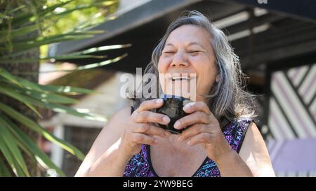 allegra donna latina anziana che beve caffè o tè all'aperto nelle soleggiate giornate estive ridendo Foto Stock