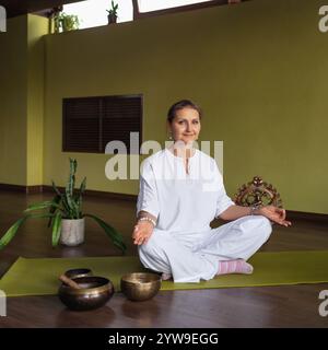 Donna che pratica kundalini yoga, meditando, seduta in posizione di loto su un tappetino in una stanza in abiti bianchi, facendo Jnana mudra con le mani, un gestu Foto Stock