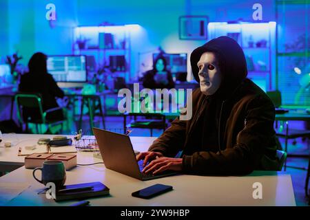 Medio primo piano di hacker che indossa felpa nera e maschera bianca che nasconde la sua programmazione del viso in ufficio buio illumato di luci al neon Foto Stock