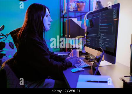 Immagine laterale di una hacker seduta sulla scrivania che guarda il monitor mentre scrive i codici sulla tastiera, indossa una maschera bianca e una felpa con cappuccio nera Foto Stock