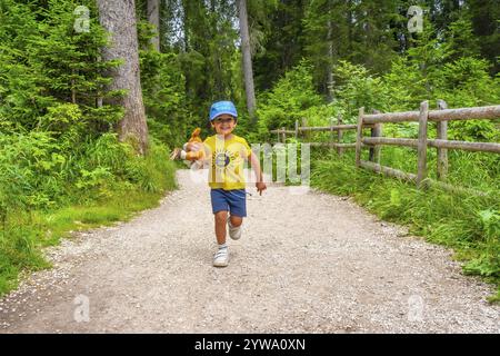 Bambino spensierato che corre lungo un sentiero di ghiaia in una foresta lussureggiante, trasportando un orsacchiotto, godendo della bellezza naturale delle dolomiti italiane Foto Stock