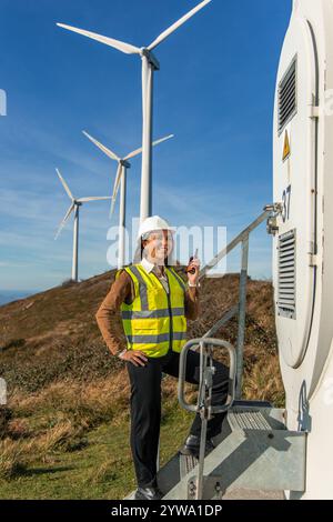 Una donna latina sorridente in un giubbotto di sicurezza e elmetto tiene un walkie-talkie mentre si trova su scale metalliche all'ingresso di una turbina eolica, con turbi Foto Stock