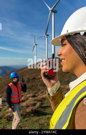 Gli ingegneri latini, donne e uomini, che indossano giubbotti di sicurezza e caschi, utilizzano walkie talkie per comunicare in un parco di turbine eoliche, lavorando su ener verde Foto Stock