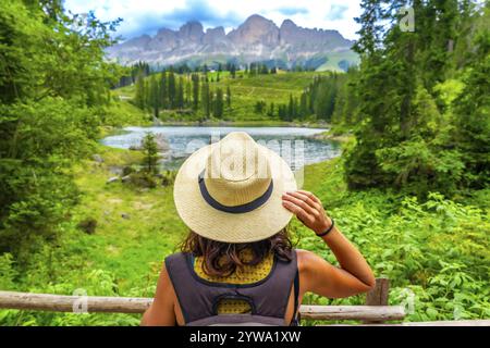 Turista donna che indossa un cappello di paglia che gode del paesaggio del lago di carezza con la catena montuosa del latemar sullo sfondo delle dolomiti in una giornata estiva Foto Stock