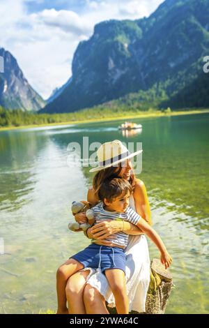Madre che abbraccia suo figlio seduto su un ceppo d'albero sulla riva del lago dobbiaco, circondato dallo splendido scenario delle dolomiti Foto Stock