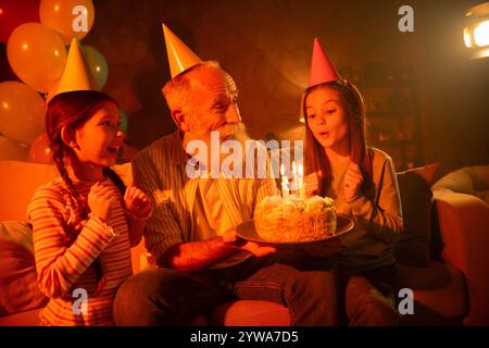 Nonno che festeggia il compleanno con le nipoti nell'accogliente soggiorno, indossa cappelli da festa, gusta torte e candele Foto Stock