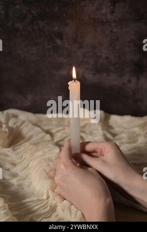 Una candela bianca illuminata con le mani di una donna su sfondo grigio scuro, panno beige Foto Stock