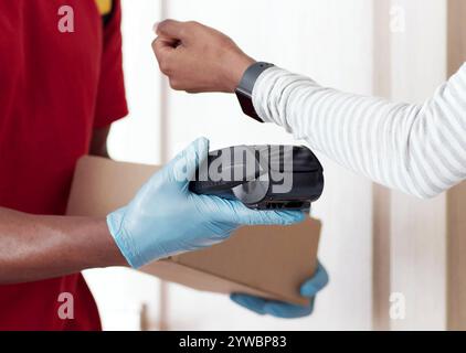 Primo piano di una donna nera che paga l'ordine con uno smartwatch, che prende la scatola dal corriere con guanti protettivi Foto Stock
