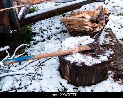 L'ascia giace su un ceppo di legno con una pila di legna da ardere tritata sul terreno innevato. Riscaldare la casa con il legno. Villaggio in Transcarpathia Ukra Foto Stock