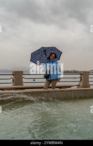 Attraente donna di mezza età in piedi sotto la pioggia di fronte a una fontana sul lungolago di Stresa Foto Stock