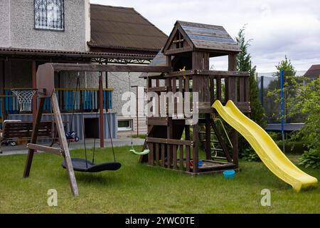 Parco giochi in legno sul retro con scivolo giallo, altalene e canestro da basket in un giardino suburbano in una giornata nuvolosa Foto Stock