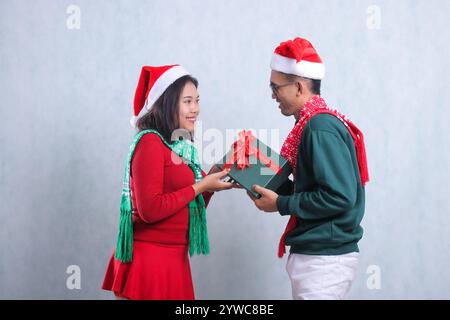 ritratto di una giovane coppia che indossa maglioni natalizi, cappelli di babbo natale e sciarpe, che si guardano allegramente l'un l'altro, fidanzato che regala una confezione regalo verde a gi Foto Stock