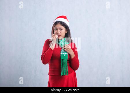 Ragazza asiatica spaventata preoccupata per la macchina fotografica che morde il dito e tiene in mano la sciarpa, indossa abiti da festa maglione di natale, cappello e sciarpa di Babbo Natale, isolata su bianco Foto Stock