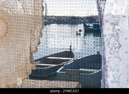 Vista delle tradizionali barche luzzu maltesi nel porto attraverso tovaglie di merletto in un mercato domenicale, Marsaxlokk, Malta Foto Stock