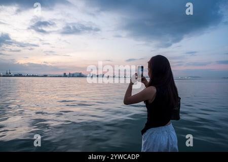 Vista posteriore di una giovane donna in piedi vicino al mare che scatta foto con il suo cellulare, Tan Jetty, George Town, Penang, Malesia Foto Stock