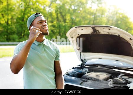 Un ragazzo nero stressato in piedi vicino a un vehice rotto, che ha una conversazione con il telefono cellulare e un servizio stradale in caso di guasto all'aperto Foto Stock