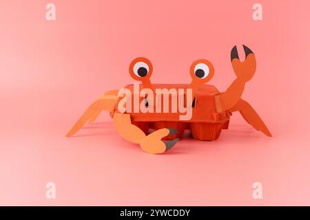 Crea un progetto che assomiglia al granchio realizzato in cartone d'uovo dipinto con gambe di carta, artigli e occhi. Artiglio ondulato e volto sorridente su sfondo rosa. Divertimento Foto Stock