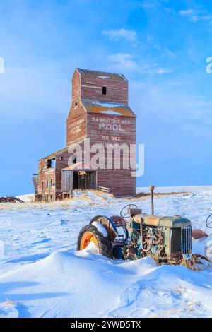 Un vecchio trattore agricolo è seduto nella neve accanto a un silo di grano. Il trattore è arrugginito e il silo è vecchio e abbandonato. Concetto di nostalgia e il Foto Stock