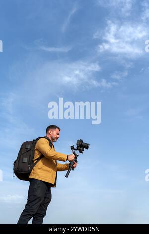 Videografo che cattura filmati con fotocamera professionale e stabilizzatore cardanico, zaino da trasporto, contro un cielo azzurro Foto Stock