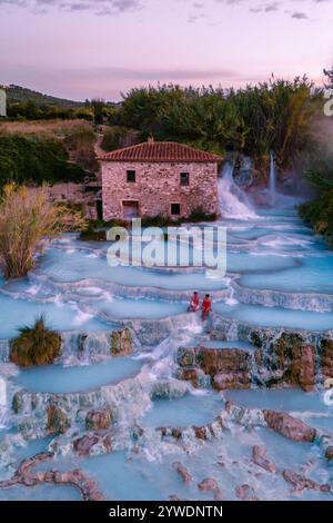 I visitatori si rilassano nelle incantevoli acque termali di Saturnia, circondati da vegetazione lussureggiante e cascate, mentre il sole tramonta, gettando un magico bagliore su questa tranquilla oasi. Foto Stock