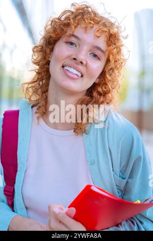 Studente dai capelli rossi sorridente con zaino e libri all'aperto. Verticale Foto Stock