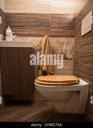 WC a parete con sedile in legno e asciugamano giallo in un bagno moderno con piastrelle marroni. Foto Stock