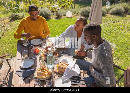 Tre amici, due uomini afroamericani e una donna, si godono un viaggio glamping in Europa, assaporando un delizioso pranzo nella natura. Foto Stock