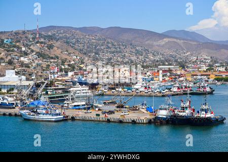 Numerosi pescherecci e rimorchiatori attraccano lungo il molo di Ensenada, Messico. Paesaggio collinare con edifici residenziali e strutture industriali in t Foto Stock