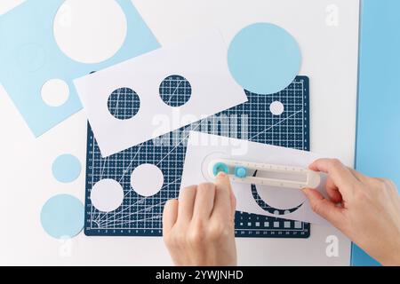 visualizzare le mani dall'alto verso il basso utilizzando lo strumento di taglio rotante manuale per tagliare il cerchio di carta in dimensioni diverse, uno sfondo blu e sono disponibili diversi circuiti di ritaglio Foto Stock