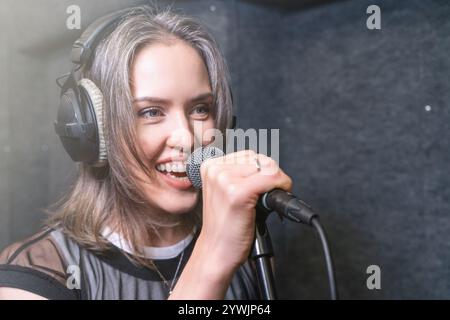 Donna che canta nel microfono in studio di registrazione, indossa le cuffie e sorride durante l'esecuzione, esprimendo gioia ed energia durante la sessione di registrazione vocale. Foto Stock