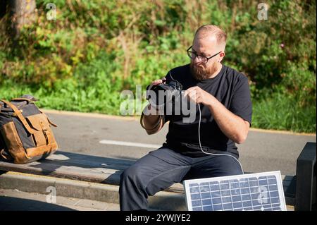 Il fotografo maschio con barba si siede sulla panca, collegando la fotocamera al pannello solare per la ricarica. Integrazione di energia rinnovabile nella fotografia, utilizzando l'energia solare per caricare le apparecchiature durante gli scatti all'aperto. Foto Stock