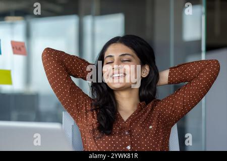 Donna d'affari sorridente che si appoggia al suo posto di lavoro, incarna relax e soddisfazione in un ambiente professionale. Mostra un momento di soddisfazione durante il lavoro e abbraccia un equilibrio Foto Stock