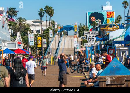 I turisti camminano lungo il molo di Santa Monica, segnando la fine della Route 66, con negozi, stand e ristoranti. Cielo soleggiato blu Foto Stock