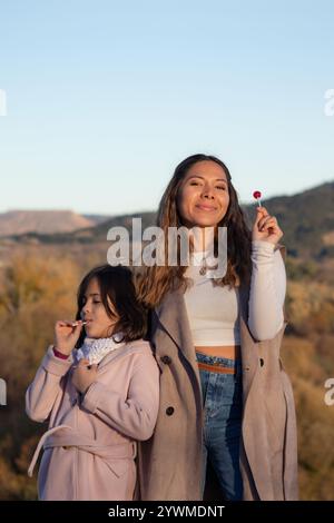Una giovane madre bruna e sua figlia si godono i lecca-lecca insieme in un paesaggio autunnale. sorrisi brillanti, irradiano felicità e gioia mentre brillano Foto Stock