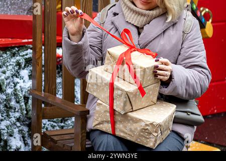 Le mani della donna sganciano il nastro rosso sui regali di Natale avvolti in carta marrone. Fuori giorno nevoso d'inverno. Primo piano. Foto Stock