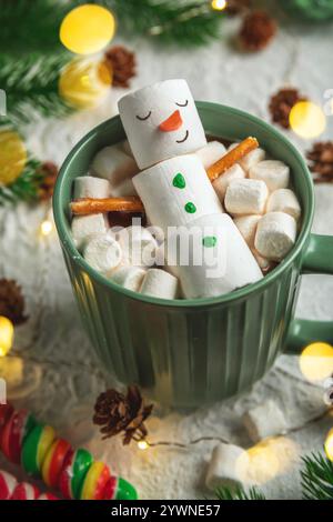 Carino biglietto di natale, allegro pupazzo di neve marshmallow si rilassa in una tazza verde di cioccolata calda, circondata da mini marshmallow, bastoncini di cannella e festi Foto Stock
