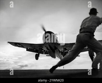 Equipaggio marittimo che assiste l'atterraggio di un Grumman F6F Hellcat sul ponte di volo del vettore. Un aereo da caccia americano basato sulla portaerei della seconda guerra mondiale, è stato progettato per sostituire il precedente F4F Wildcat e contrastare il giapponese Mitsubishi A6M Zero. L'aereo ha fatto il suo debutto in combattimento nel settembre 1943 e successivamente si è affermato come un robusto caccia portaerei ben progettato e divenne il caccia dominante della United States Navy nella seconda metà della guerra del Pacifico. In totale, 12.275 furono costruiti in poco più di due anni e alla Hellcats fu attribuito il merito di aver distrutto un totale di 5.223 aerei nemici. Foto Stock