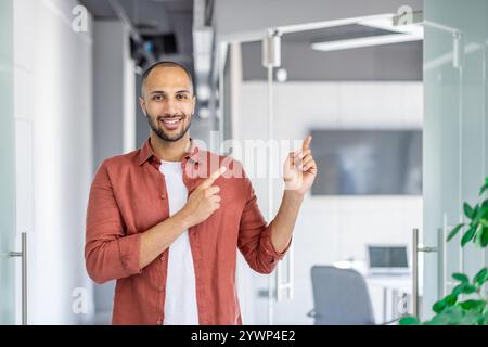 Un giovane e allegro professionista in un outfit casual punta verso una porta in uno spazio ufficio luminoso, simboleggiando la guida, la professionalità e l'entusiasmo. Foto Stock