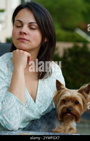 Una ragazza dai capelli scuri vestita leggermente sorridente si siede con il suo piccolo cane a un tavolo in un bar in estate. Yorkshire terrier tra le braccia del suo proprietario Foto Stock