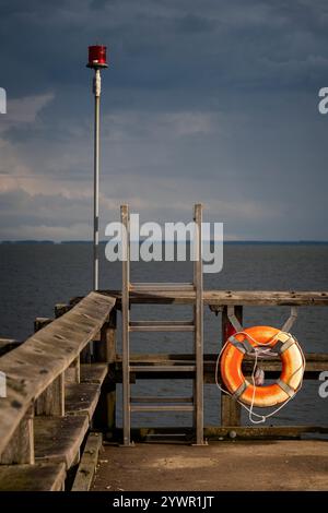 Molo in legno con anello di salvataggio, scala in metallo e luce rossa di sicurezza contro un cielo moody su un mare calmo. Foto Stock