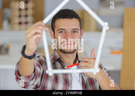 il giovane lavoratore mostra la forma della casa dal righello con i pollici in alto Foto Stock