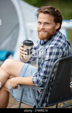 un giovane hipster barbuto siede su una sedia pieghevole in campeggio Foto Stock