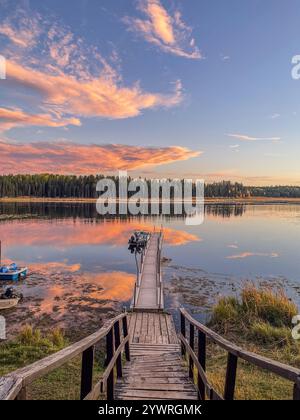 Una banchina di legno rustica che si estende in un tranquillo lago, incorniciata da un cielo vibrante al tramonto e dal riflesso speculare degli alberi e delle montagne circostanti Foto Stock