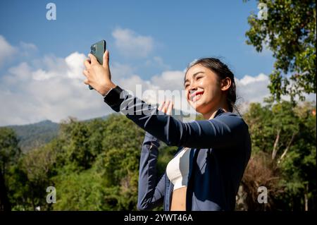 Un'allegra donna asiatica in abbigliamento sportivo scatta un selfie, registra un video o parla durante una videochiamata sul suo smartphone mentre si esercita nel parco in m Foto Stock