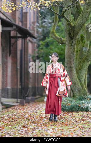 Una donna giapponese sui vent'anni cammina di fronte a un grande albero e a edifici tradizionali giapponesi, indossando un kimono rosso (hakama), un punto fermo per un giapponese Foto Stock