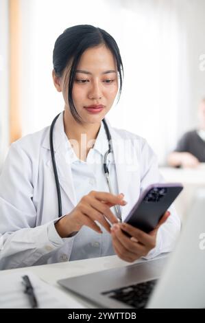 Una bella e positiva dottoressa asiatica in uniforme si siede alla sua scrivania in ufficio, leggendo del testo sul suo smartphone. persone e tecnologia wireless Foto Stock