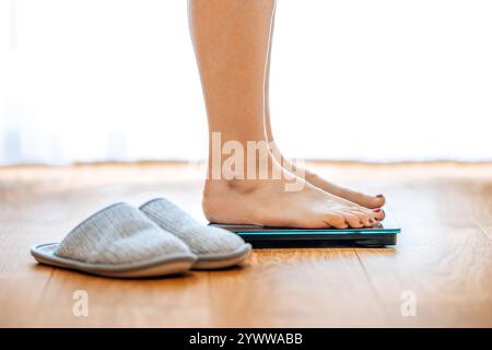 La donna sta in piedi sulla bilancia e si pesa la mattina in salotto, vista ravvicinata da un lato Foto Stock