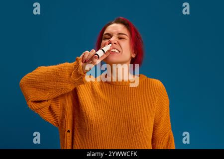 Ritratto di giovane donna si sente male a causa di starnuti e spruzzare il naso per rimuovere i sintomi su sfondo blu scuro. Foto Stock