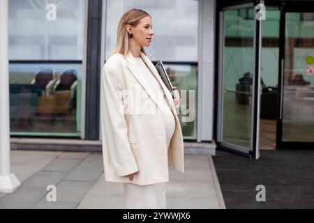 Una bella donna bionda incinta con un abito da lavoro bianco e un blazer si trova all'aperto con un notebook vicino a un edificio aziendale, che mostra una professionista Foto Stock